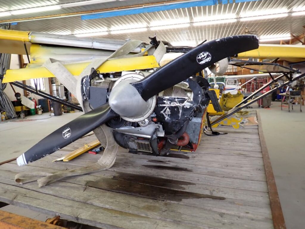 A close-up view of the damaged nose, engine, and propeller of the yellow Avid Magnum wreckage inside a hangar. The propeller blades are bent and chipped, and the engine compartment shows deformation and impact damage.